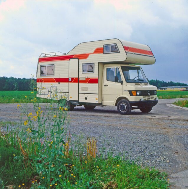 1980_Alkoven-Fahrzeug mit rotem Streifendesign – geparkt auf Asphaltfläche mit Blumen im Vordergrund.