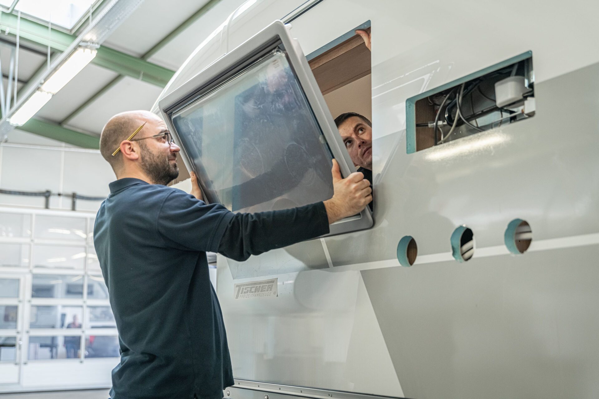 Zwei Mitarbeiter der Tischer Manufaktur setzen gemeinsam ein Fenster in die Seitenwand einer Wohnkabine ein.