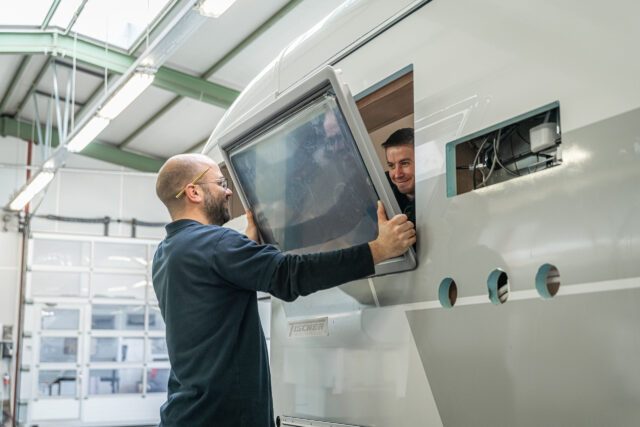 Zwei Mitarbeiter der Tischer Manufaktur setzen gemeinsam ein Fenster in die Seitenwand einer Wohnkabine ein.
