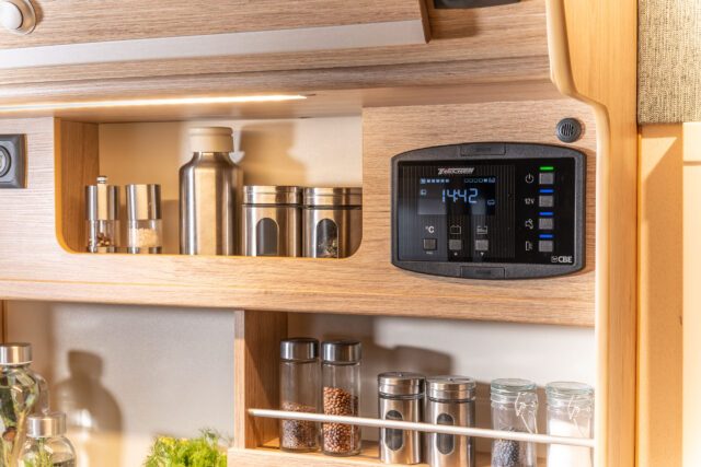 Bedienelement mit Display in einem Regal zwischen Edelstahlflaschen und Gewürzgläsern im Küchenbereich.