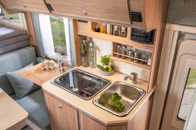 Trail230S Kompakte Küchenzeile mit Spüle, Kochfeld und Stauraum in Tischer Wohnkabine – mit Blick nach draußen.