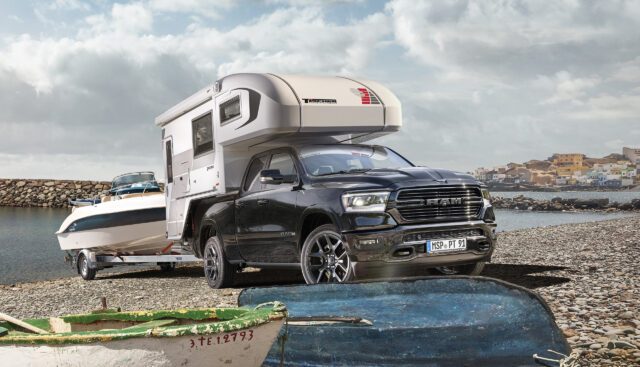 RAM Pickup mit Tischer-Kabine und Bootsanhänger auf Kiesstrand vor Hafenstadt.