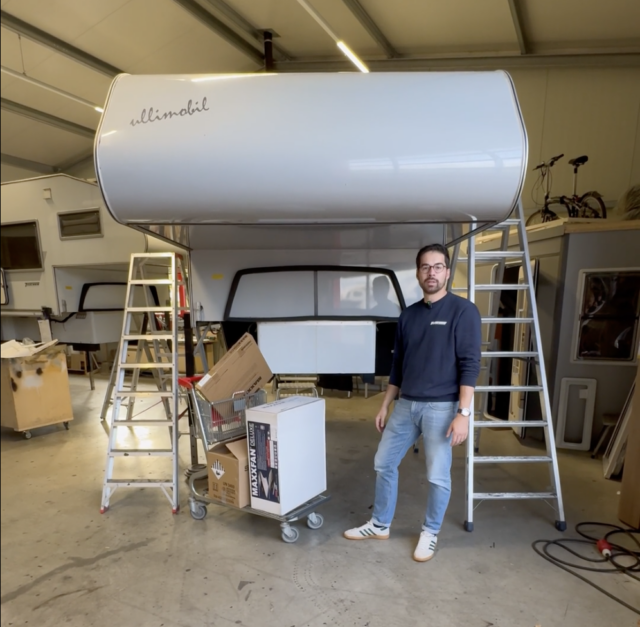 Unmontierte Wohnkabine in Werkstatt mit Leitern, Mitarbeiter zeigt Aufbau.