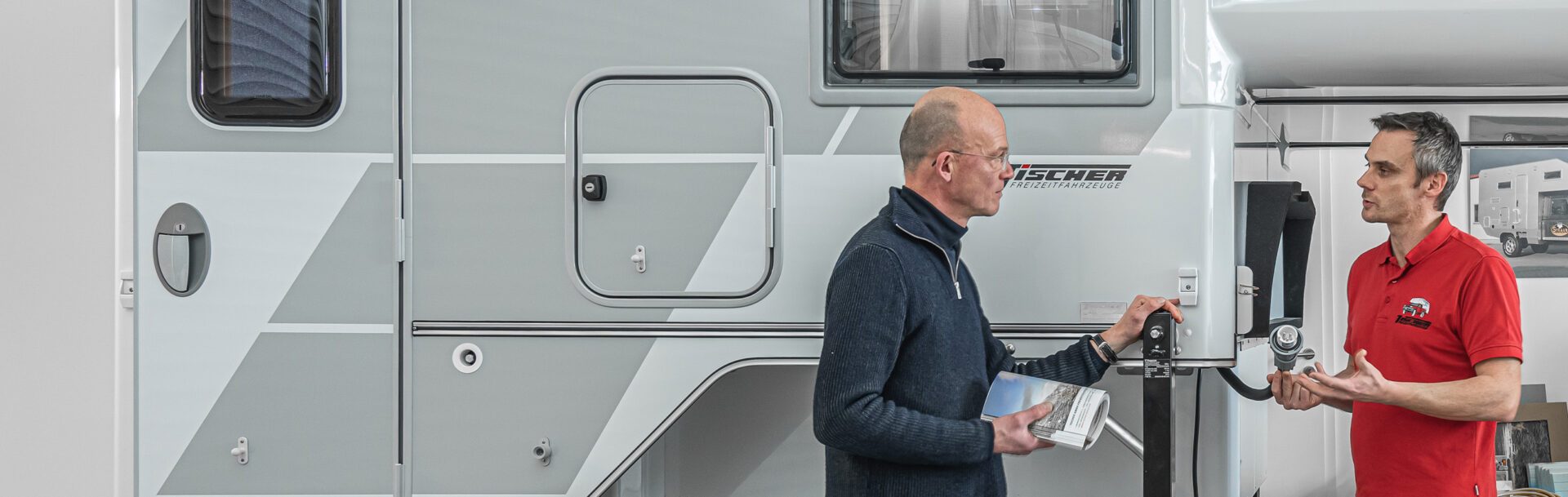 Zwei Mönner vor der Seitenwand einer Tischer-Wohnkabine mit klar erkennbarer Tür und grafischen Elementen.