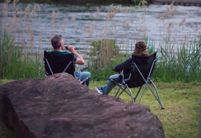 Zwei Personen sitzen in Campingstühlen am Wasser und genießen den Ausblick – entspannte Szene im Tischer-Urlaub. MIetfahrzeug